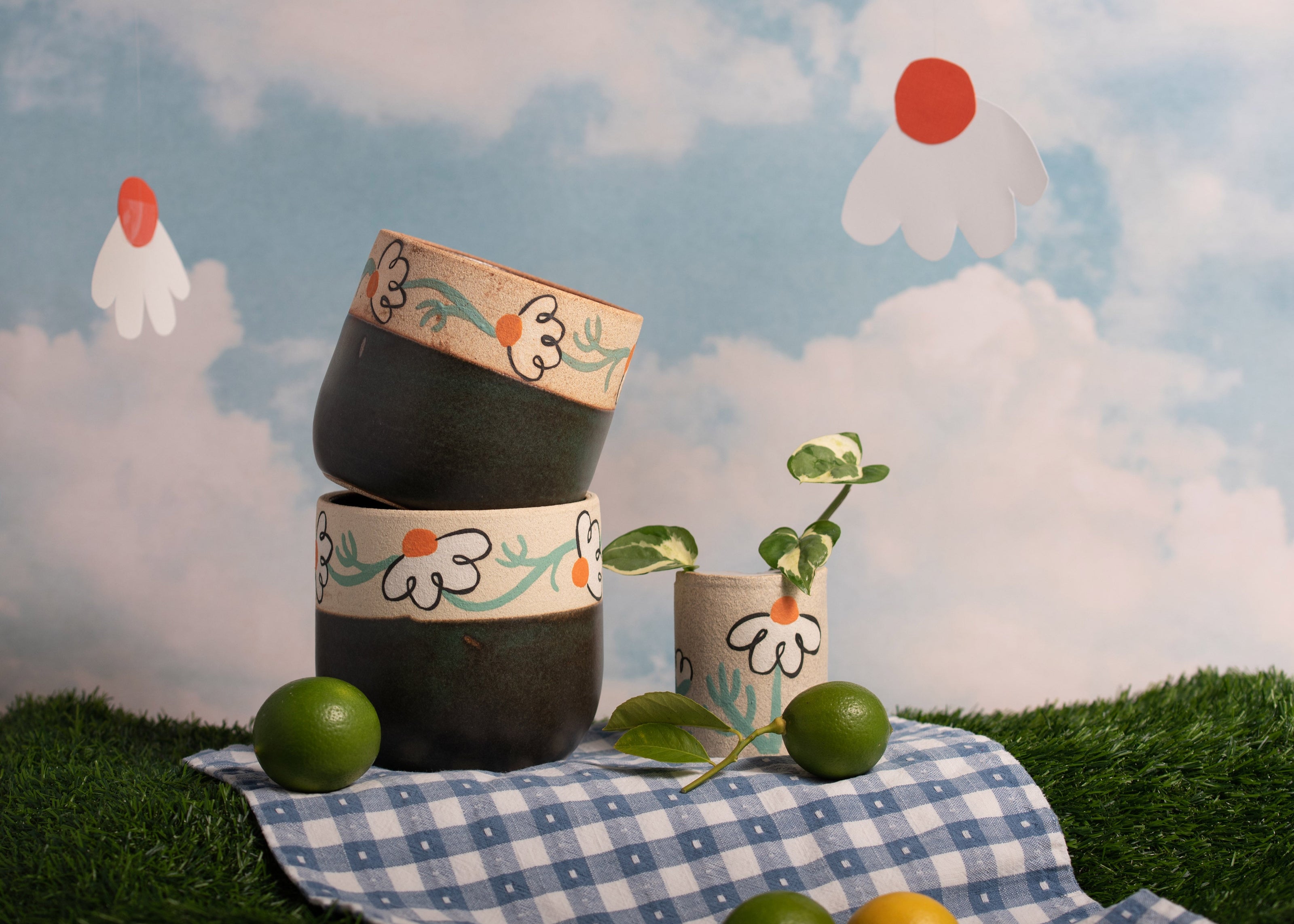 Decorative ceramic pots with floral patterns on a checkered cloth against a whimsical sky background.
